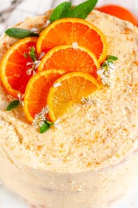 Orange Cake with Zesty Cream Cheese Frosting with orange slices and sage leaves on white cake stand over head close up