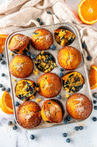 Zesty Orange Blueberry Muffins in silver muffin tray with sliced oranges and fresh blueberries on white marble overhead view