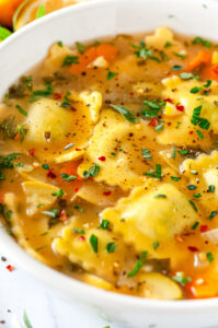 One Pot Ravioli Soup in white bowl with lemons and sage on white marble