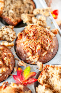 Maple banana muffins in gray metal muffin pan with walnuts and leaves on white marble