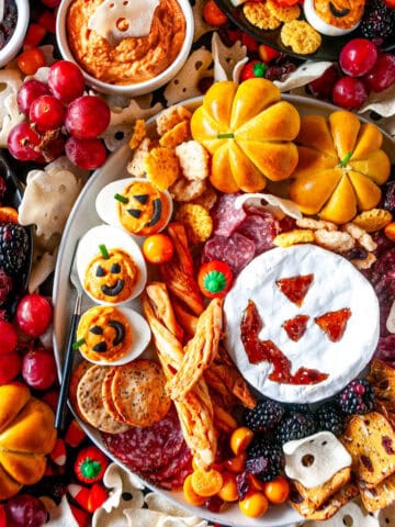 Top-down view of a Halloween cheese board filled with spooky snacks, including a Jack-o'-lantern brie, ghost-shaped chips, cured meats, mini pumpkins, roasted red bell pepper hummus, and Halloween deviled eggs.